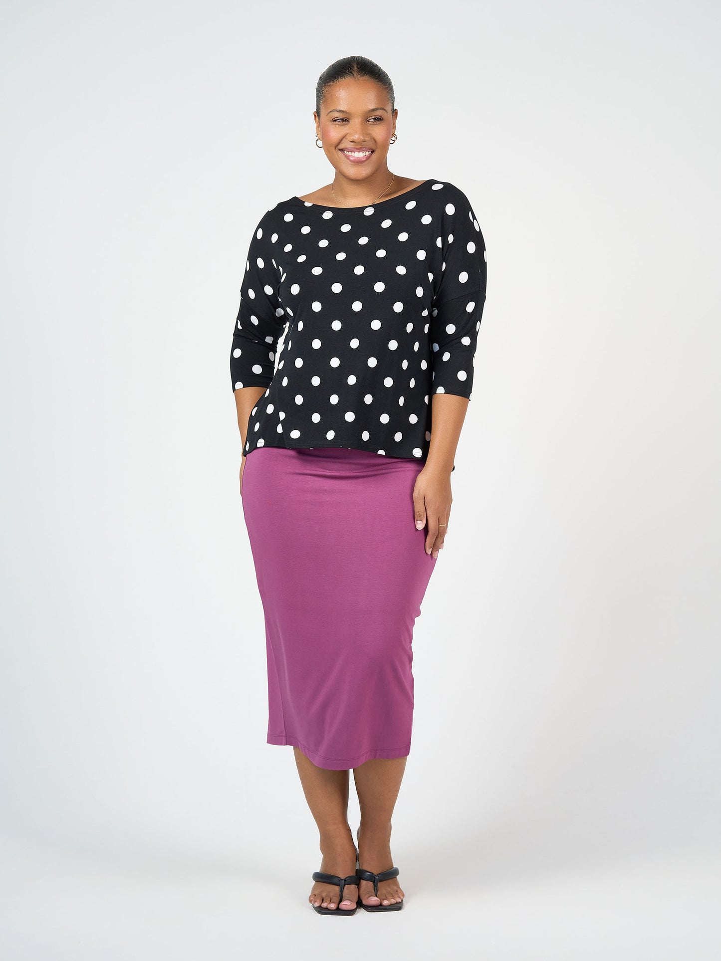 A woman smiles, wearing the Camila 3/4 Sleeve Bamboo Top in black and white polka dot with a purple midi skirt, her hair tied back and paired with black sandals against a plain white background.