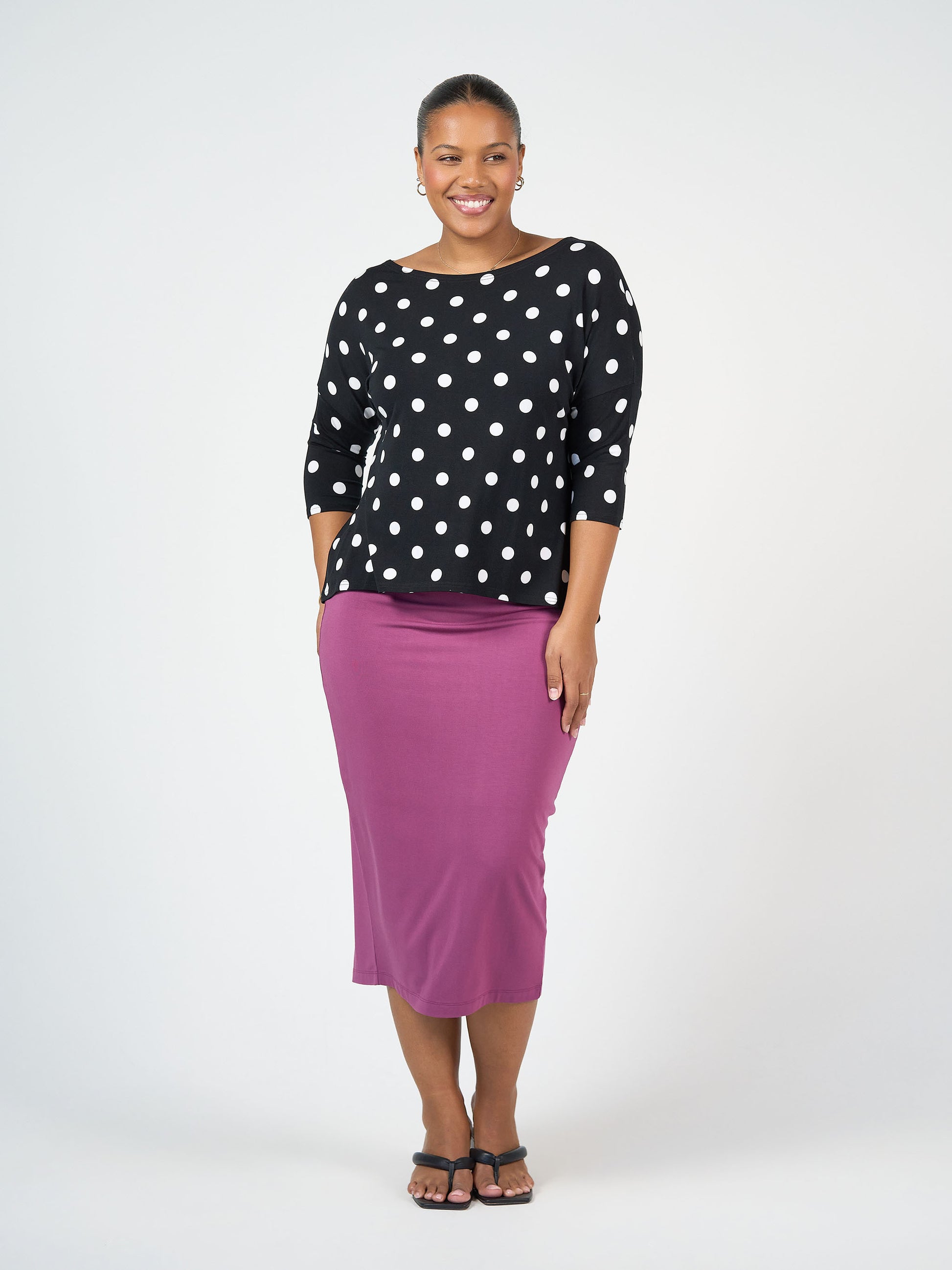 A woman smiles, wearing the Camila 3/4 Sleeve Bamboo Top in black and white polka dot with a purple midi skirt, her hair tied back and paired with black sandals against a plain white background.