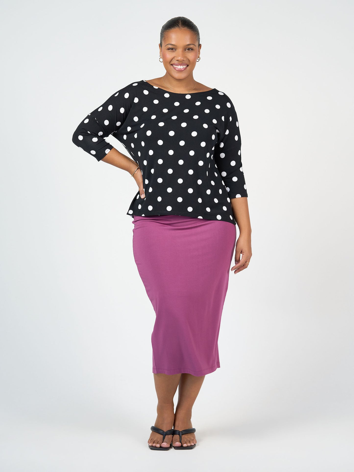 A smiling woman with one hand on her hip wears the Camila 3/4 Sleeve Bamboo Top - Polka Dot, paired with a purple midi skirt and black sandals, standing against a plain light background.