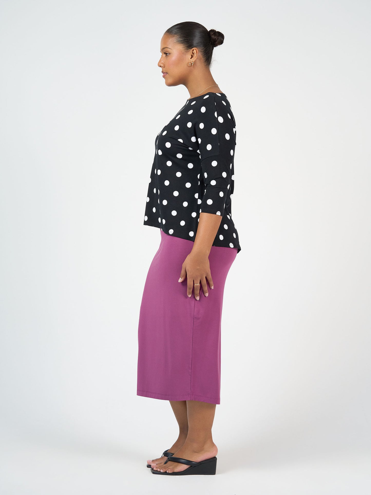 A woman stands in profile against a plain background, wearing the Camila 3/4 Sleeve Bamboo Top - Polka Dot, a long magenta skirt, black wedge sandals, and her hair styled in a neat bun.