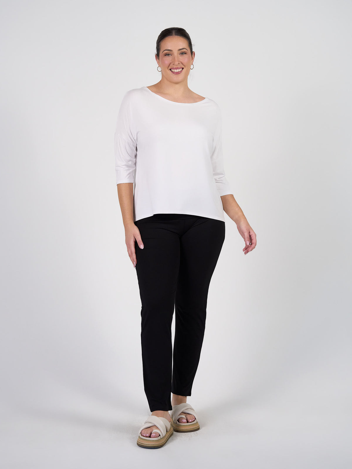 A woman stands against a white background, smiling and wearing the Camila 3/4 Sleeve Bamboo Top in White with black pants and beige sandals. Her hair is pulled back, and she rests one hand by her side.