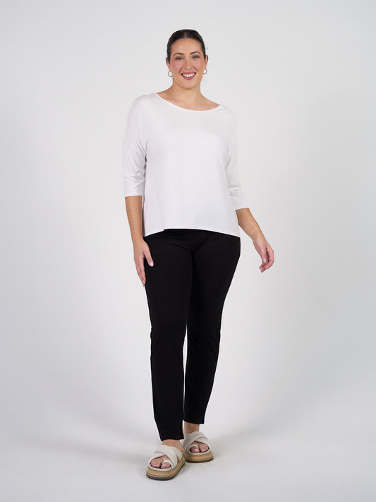 A woman stands against a white background, smiling and wearing the Camila 3/4 Sleeve Bamboo Top in White with black pants and beige sandals. Her hair is pulled back, and she rests one hand by her side.