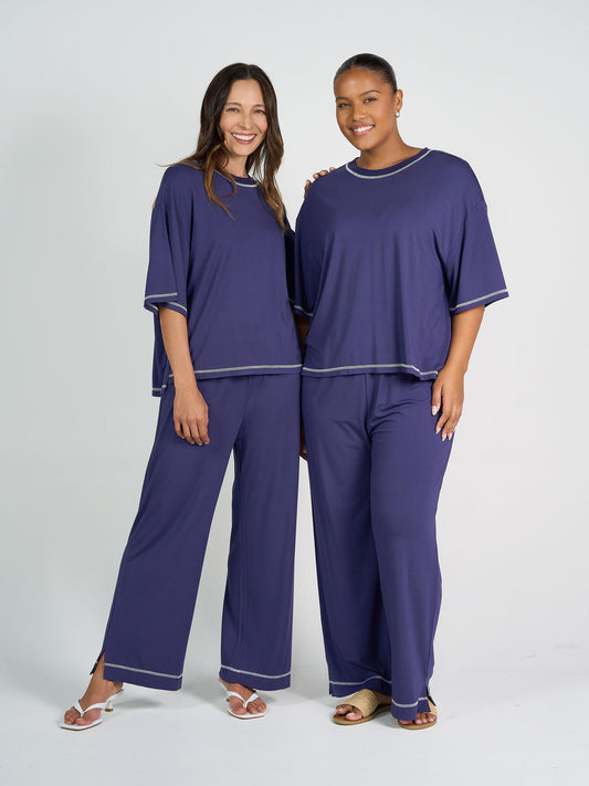 Two women smile side by side in matching dark purple Carey Bamboo Culotte loungewear, featuring ultra-soft fabric, short-sleeved tops, and wide-leg pants with silver trim. The background is plain and light-colored.