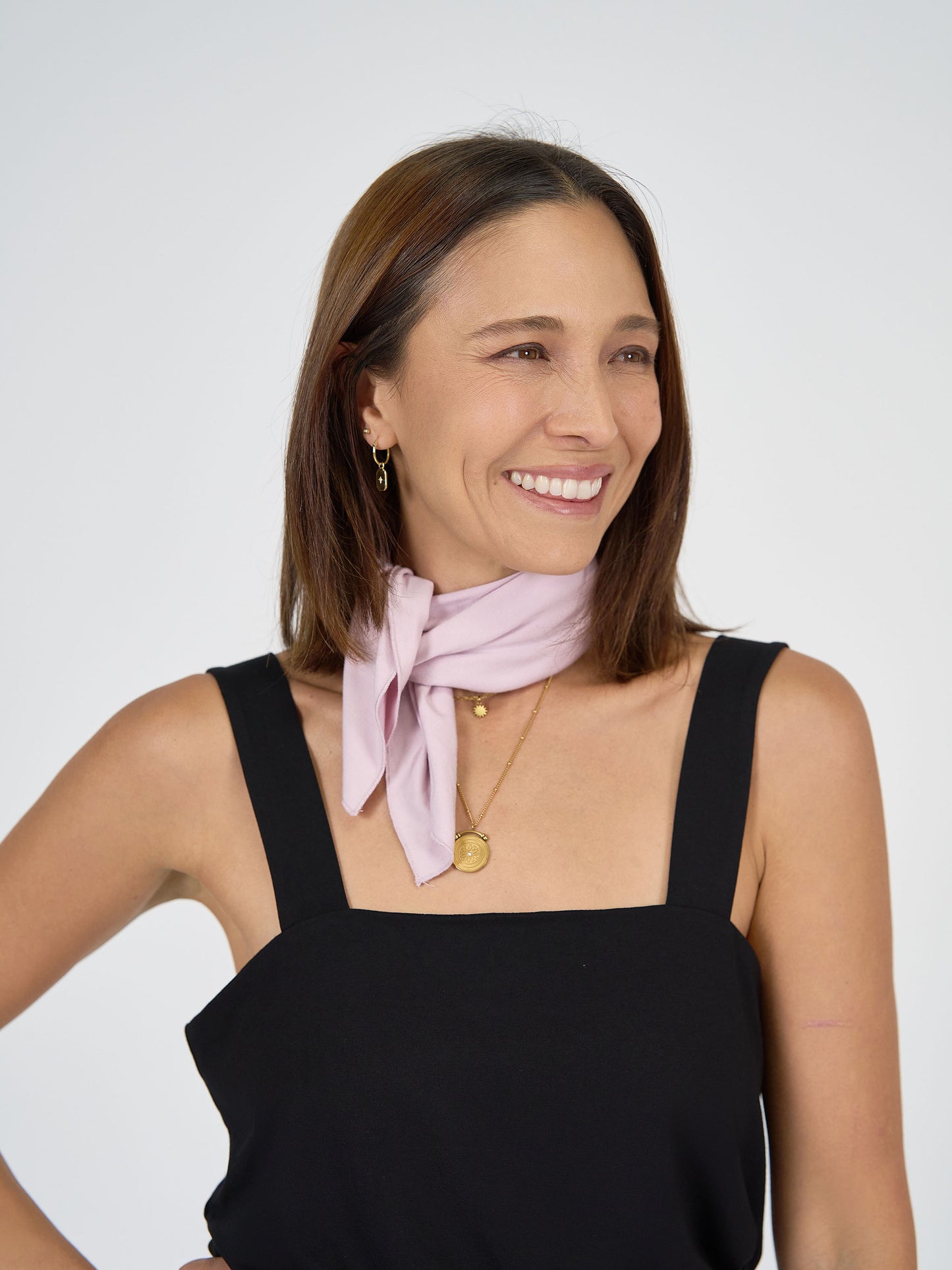 A woman with straight brown hair wears a sleeveless black top, gold jewelry, and the Benni Bamboo Scarf in Lilac draped around her neck, her smile bright against a plain light background.