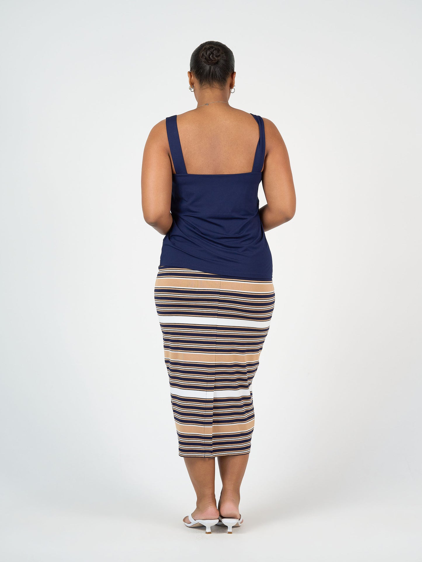 A person faces away from the camera, wearing a navy tank top, the Maxi Bamboo Whitney Tube Skirt - Stripe in tan, white, and navy, and white heeled sandals against a plain white background.