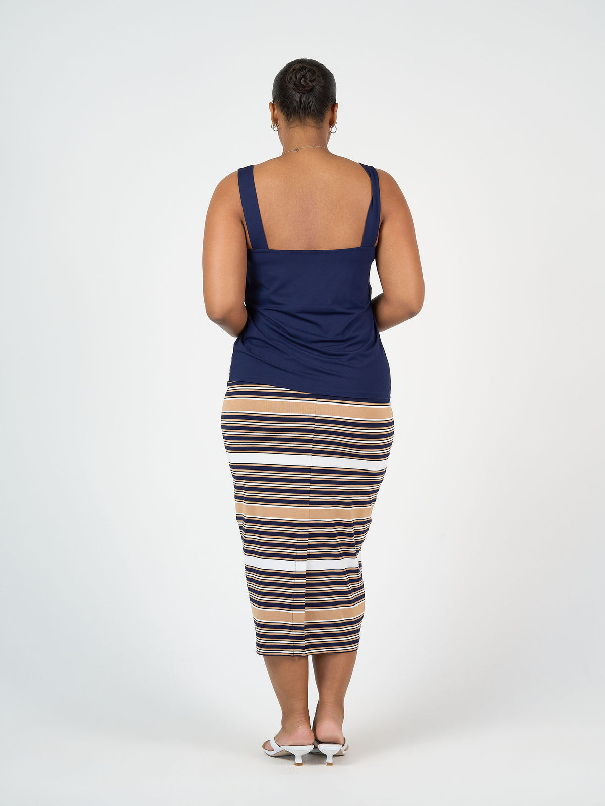A person faces away from the camera, wearing a navy tank top, the Maxi Bamboo Whitney Tube Skirt - Stripe in tan, white, and navy, and white heeled sandals against a plain white background.