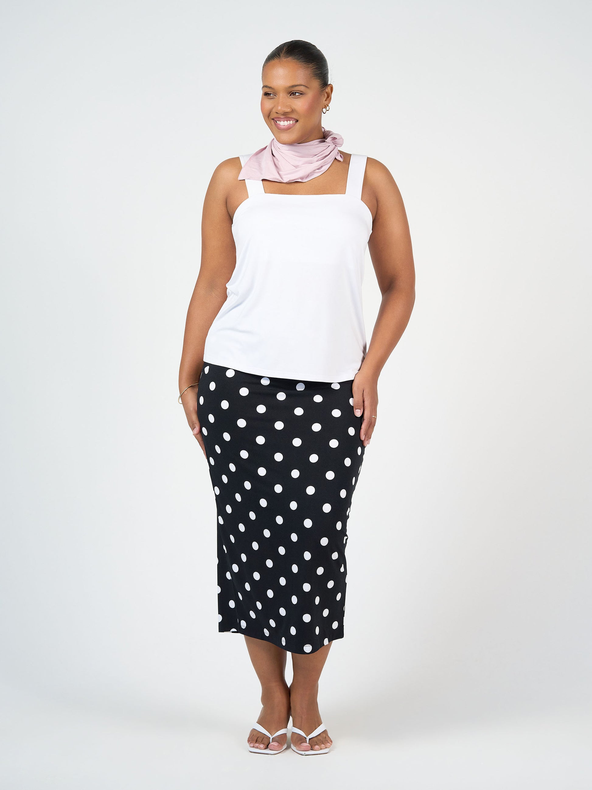 A woman smiles while wearing a white tank top, pale pink scarf, white sandals, and the Maxi Bamboo Whitney Tube Skirt - Polka Dot, standing against a plain light background.