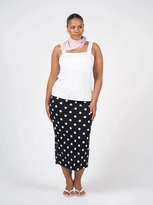 A woman stands against a white background wearing a white sleeveless top, black polka dot midi skirt, white sandals, and the Benni Bamboo Scarf - Lilac—a sustainable accessory—styled around her neck.