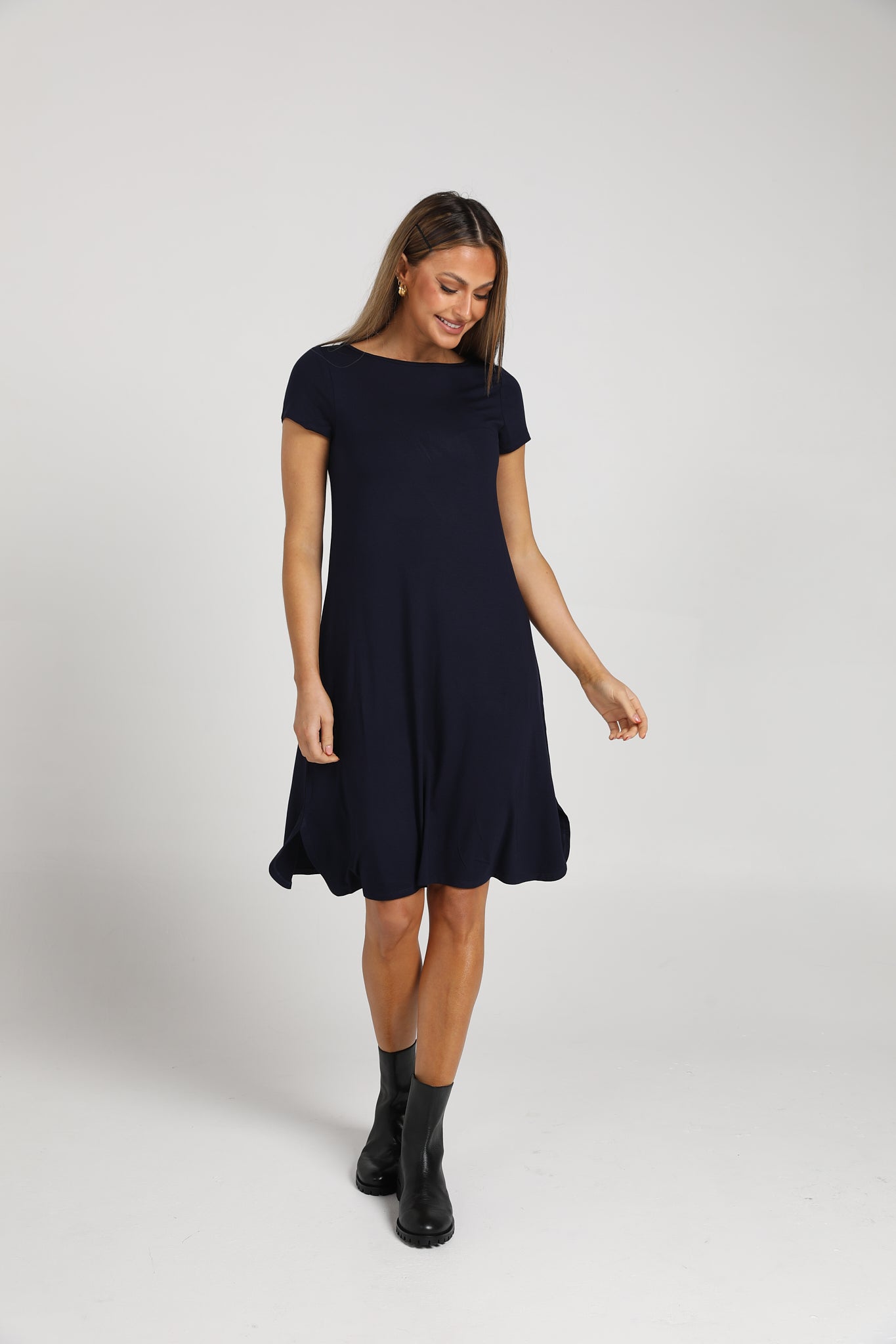 A woman in the Cher Bamboo Short Sleeve T-Shirt Dress and black ankle boots stands against a white background, smiling gently—an ideal choice for a sustainable wardrobe.