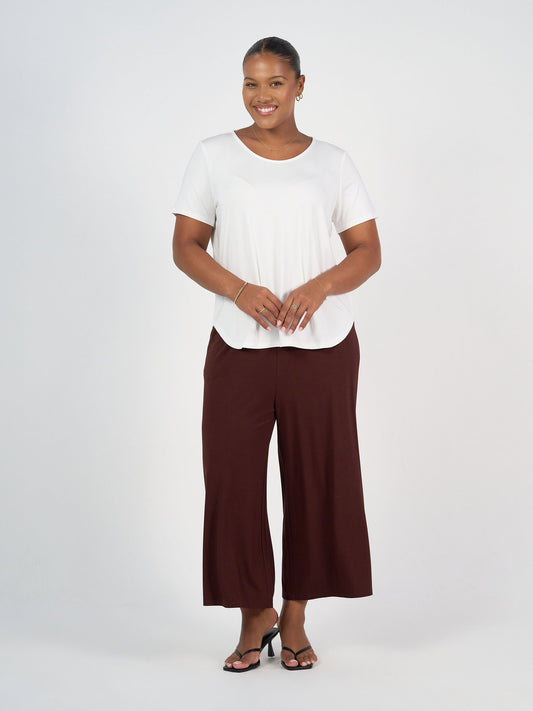 A woman stands smiling against a white background, wearing a white short-sleeve top, the Ciara 7/8 Length Bamboo Culotte Pant in Burgundy, black heeled sandals, and simple jewelry. Her hands are clasped in front of her.