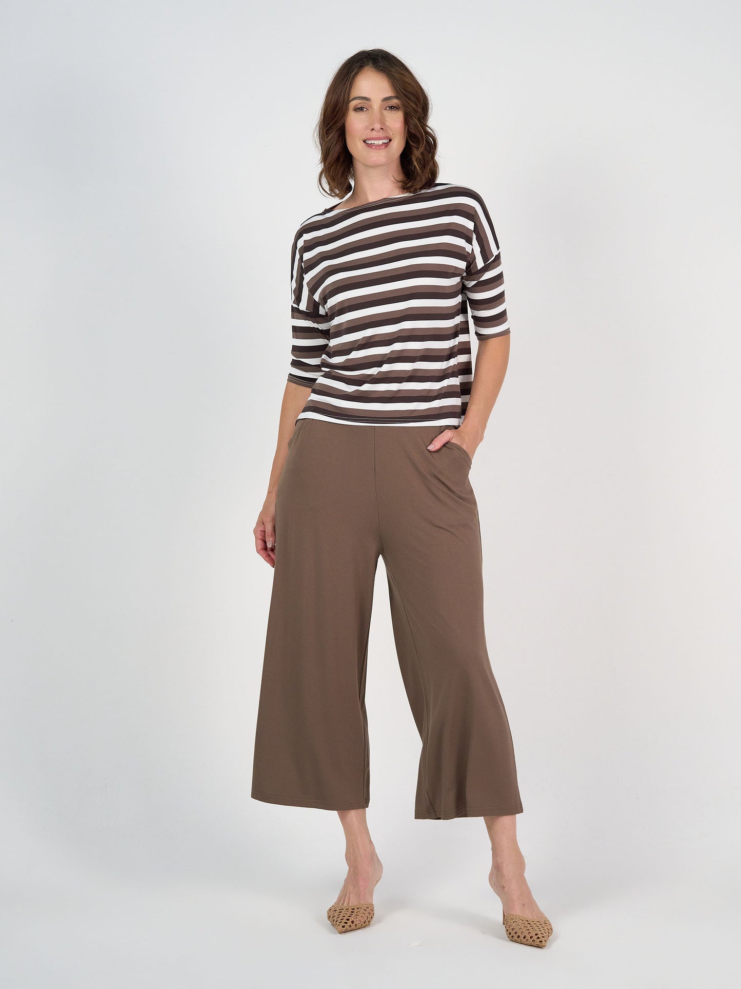 A woman stands against a plain white background, smiling with one hand in her pocket, wearing a brown and white striped top, beige woven flats, and the Ciara 7/8 Length Bamboo Culotte Pant in Chocolate.