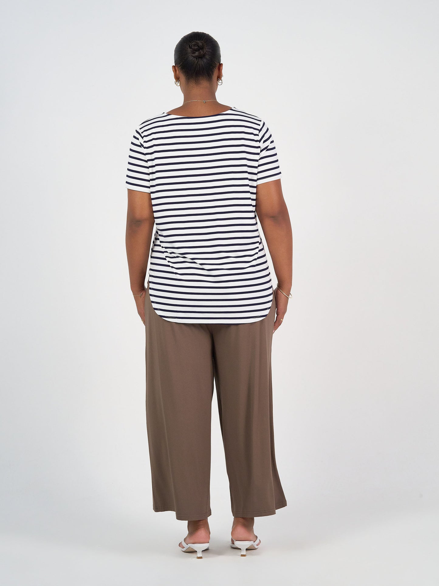 A person stands facing away from the camera in a black-and-white striped t-shirt, Ciara 7/8 Length Bamboo Culotte Pant in Chocolate, and white sandals against a plain white background.