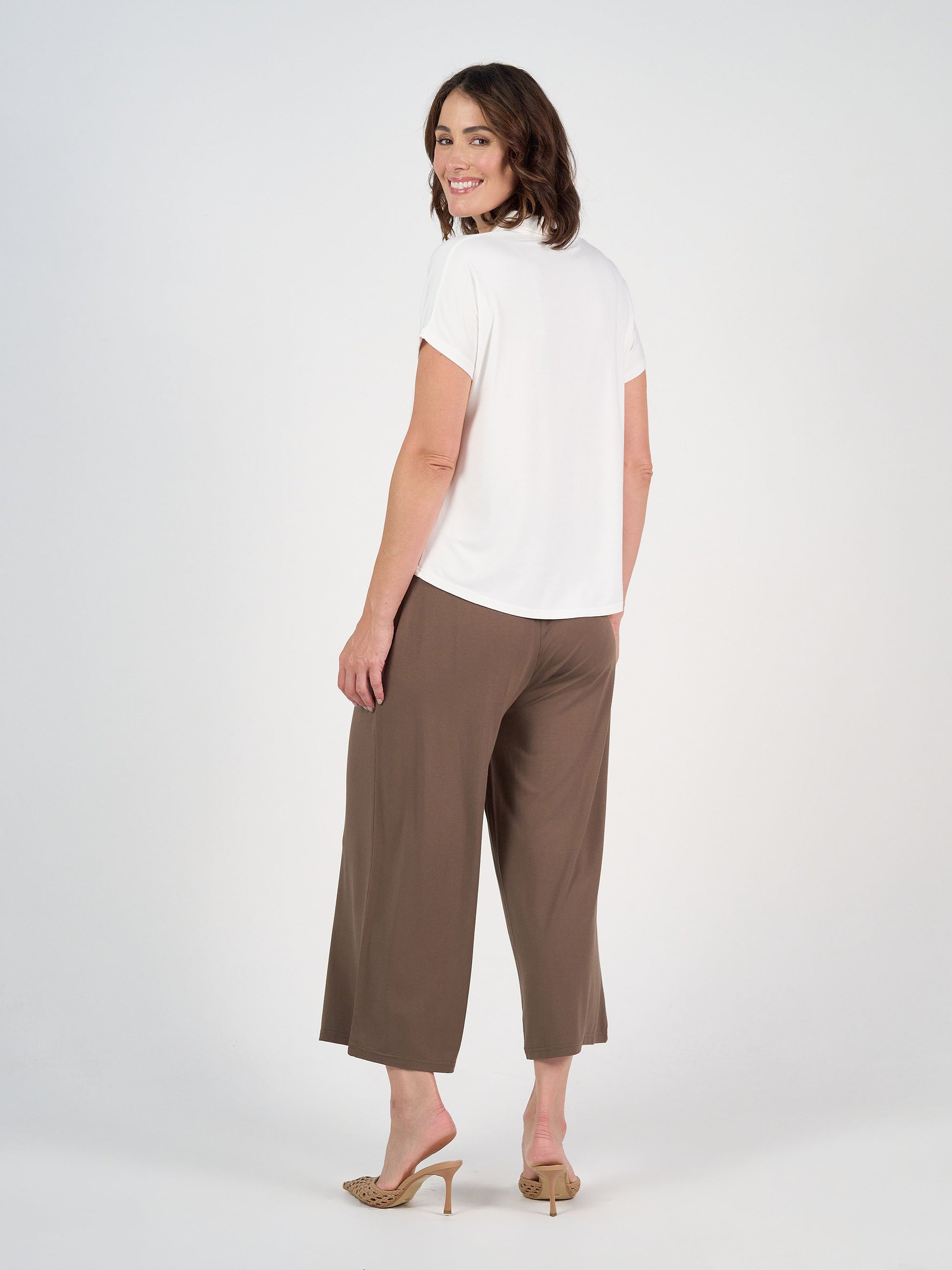 Smiling woman stands against a plain background, looking over her shoulder, wearing a white short-sleeve top and the Ciara 7/8 Length Bamboo Culotte Pant in Chocolate with beige heeled sandals.