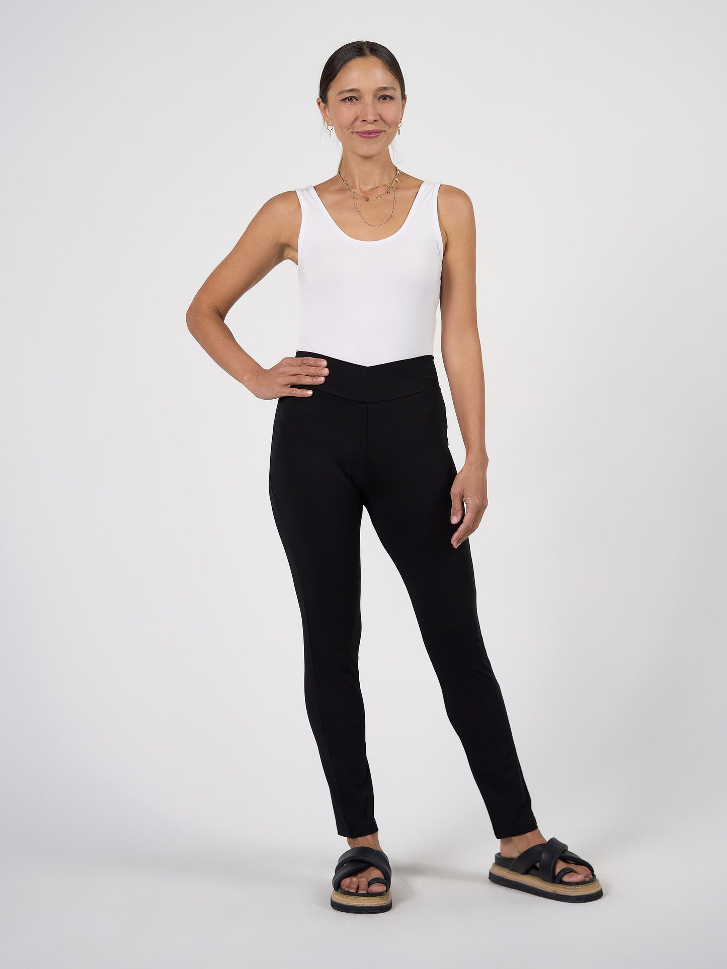 A woman stands against a white background, smiling with one hand on her hip. She wears high-waisted Patti Bamboo Full-Length Pants in black, a white sleeveless top, black sandals, and layered necklaces.