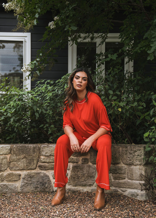 A woman wears the Carey Bamboo Culotte - Fiery Red with an elastic waistband, sitting on a stone ledge amid green foliage. With long wavy hair and tan ankle boots, she confidently gazes at the camera, a black house behind her.