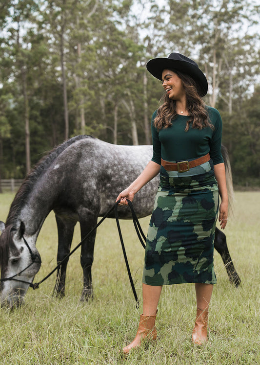 A smiling woman in a wide-brimmed black hat and the Maxi Bamboo Whitney Tube Skirt - Camouflage stands in a grassy field, holding the reins of a grazing gray horse with tall trees in the background.