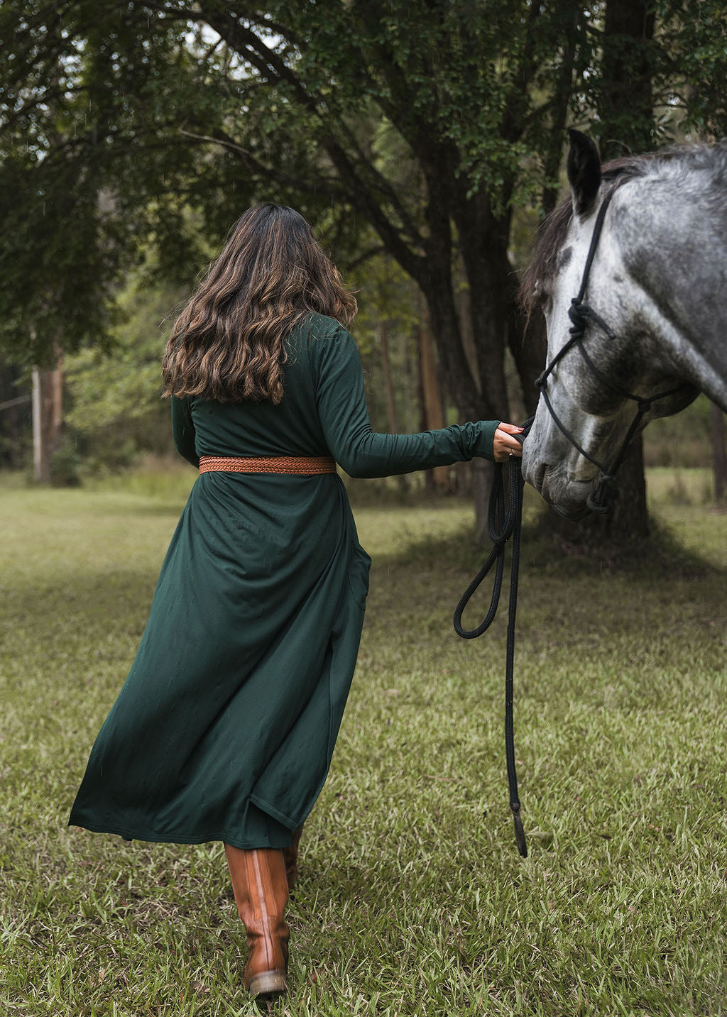 A woman with long brown hair, in a green dress, brown boots, and the Bamboo Longline Pocket Cardigan - Dark Green, walks through grass holding a gray horse’s reins. Trees can be seen in the background.
