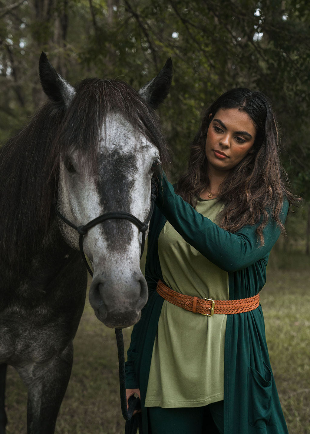 A woman with long dark hair gently pets a gray horse outdoors, surrounded by trees and grass. She wears the Bamboo Longline Pocket Cardigan in Dark Green and a brown braided belt, gazing calmly at the horse.