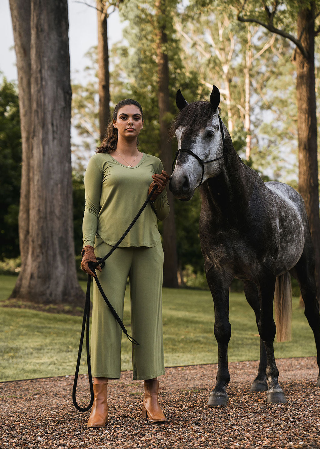 A woman wearing the Sia V Neck Bamboo Longsleeve Top in Olive Green stands outdoors on gravel, holding the reins of a gray horse. She pairs it with brown boots and gloves, with tall trees and green grass in the background.