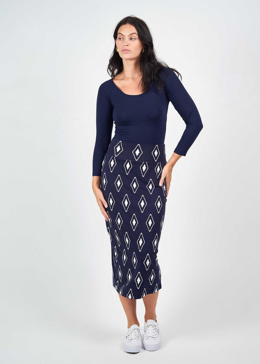A woman with dark hair and a neutral expression wears the Ava 3/4 Sleeve Cropped Bamboo Top in navy, paired with a navy midi skirt featuring a white diamond pattern and white sneakers, standing against a white background.