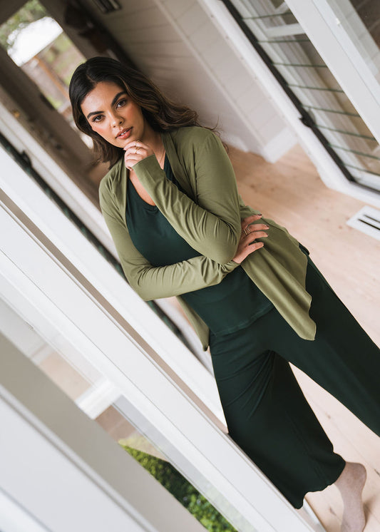 A woman with long dark hair leans against a white doorway, wearing the Joni Bamboo Waterfall Cardigan/Jacket in Olive over a dark green top and matching pants, gazing at the camera with one hand thoughtfully on her chin.