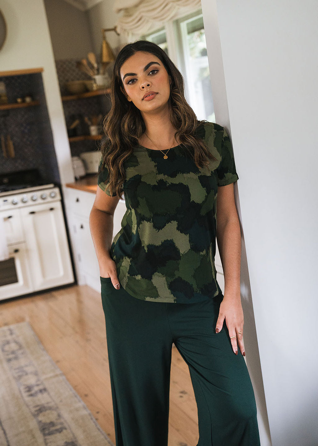 A woman with long wavy hair stands indoors in a kitchen, wearing the Janis Short Sleeve Bamboo T-Shirt in camouflage print and dark green pants, with one hand in her pocket and the other resting on a white wall.