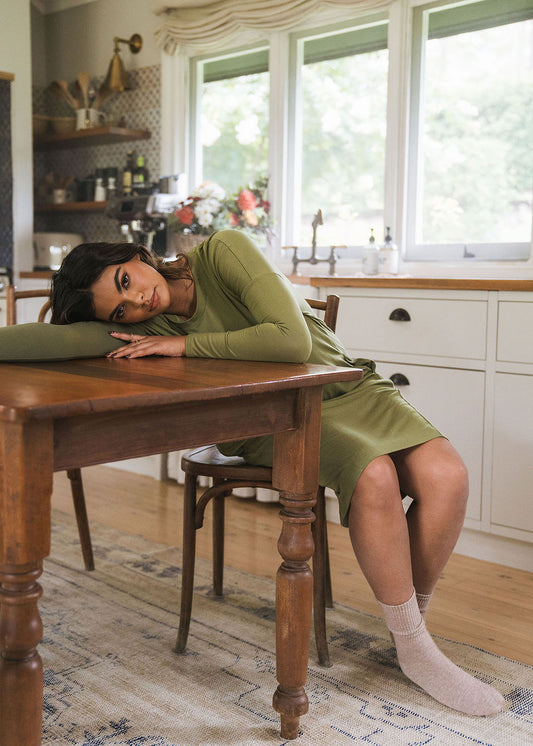 A woman in an olive Turner Long-Sleeve Bamboo T-Shirt Dress and cream socks sits at a wooden table in a bright kitchen, resting her head and arm on the tabletop while gazing thoughtfully toward the camera.