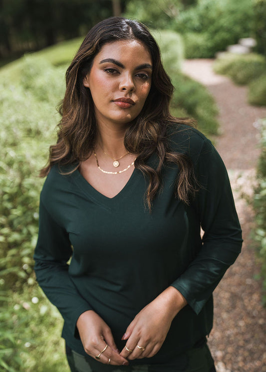 A woman with wavy brown hair wears the Braxton Bamboo Long Sleeve Top in dark green outdoors on a gravel path, accessorized with gold necklaces, and looks confidently at the camera amid lush greenery.