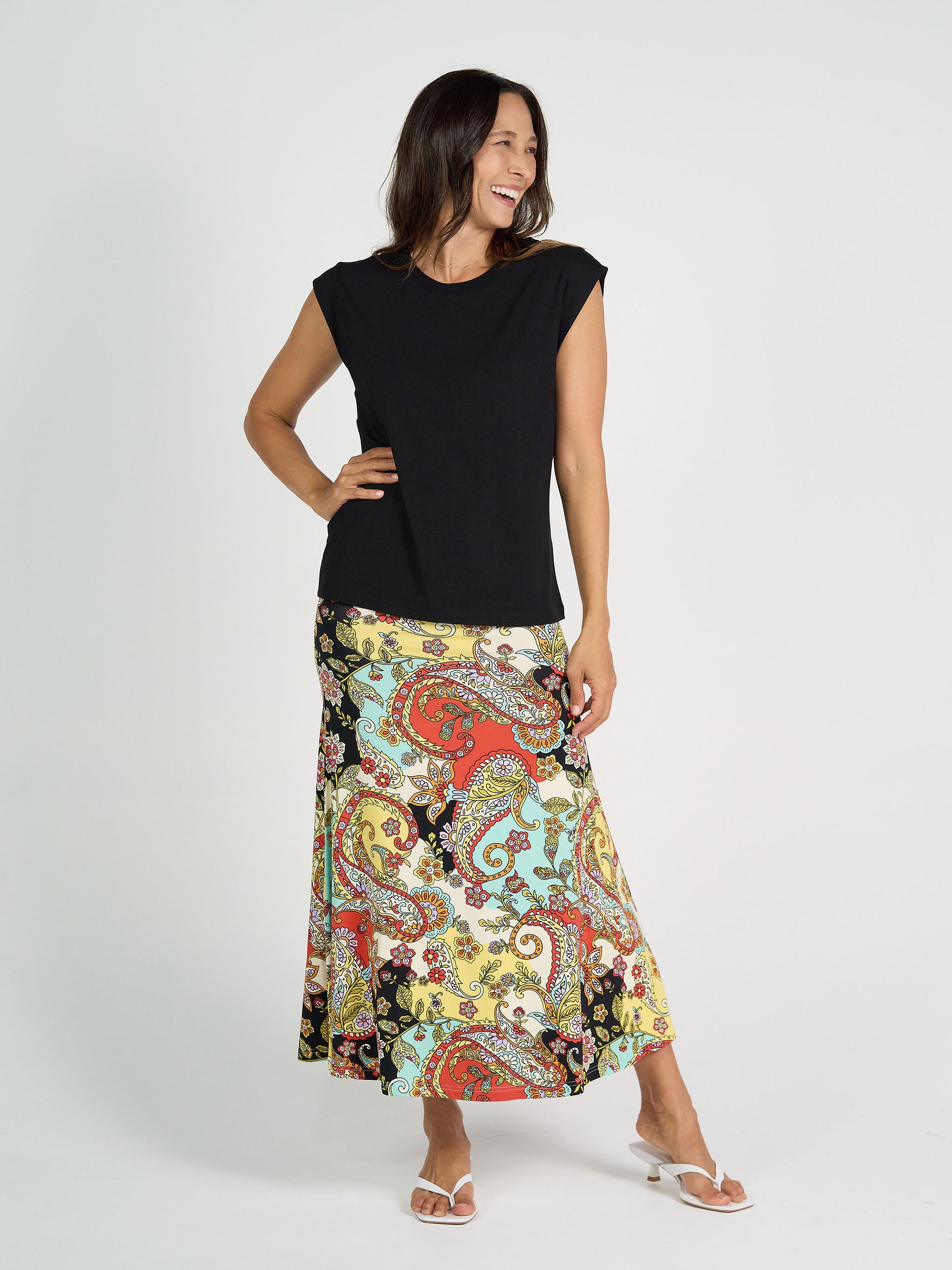 A woman smiles in the Donna Cap Sleeve Bamboo Tee and a colorful paisley maxi skirt with white sandals. She has long brown hair and poses against a plain white background.