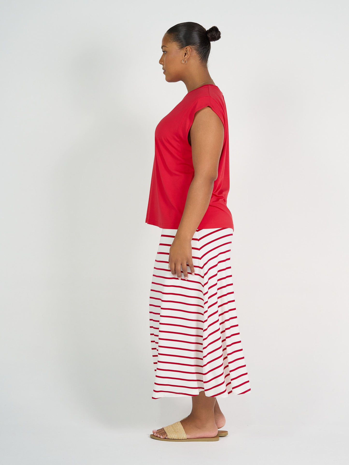 A woman stands in profile against a white background, wearing the Donna Cap Sleeve Bamboo Tee in red, a white ankle-length skirt with red stripes, flat woven sandals, and her hair tied back in a neat bun.