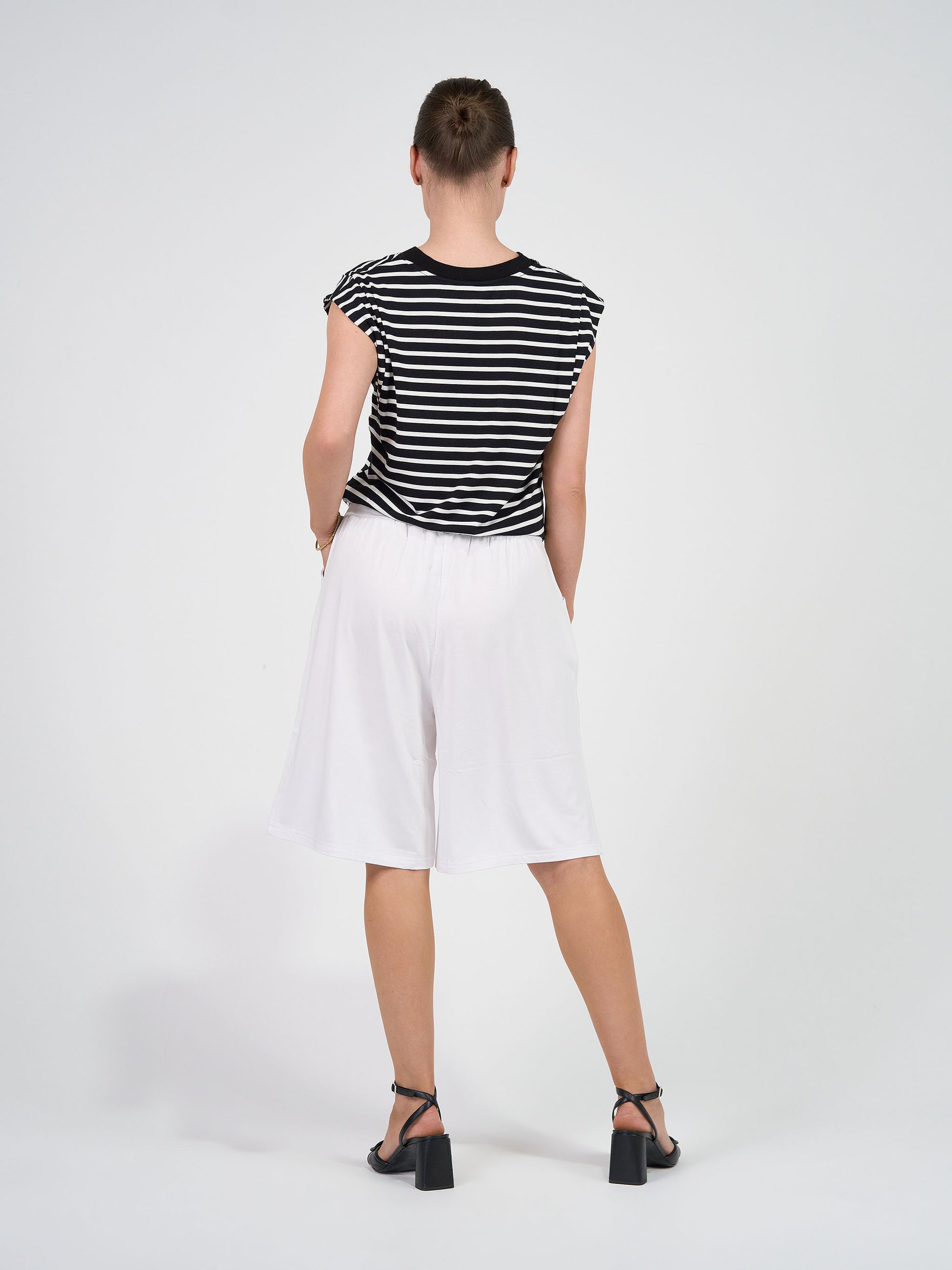 Facing away from the camera against a plain background, the person wears the Donna Cap Sleeve Bamboo Tee in black & white stripe, paired with loose white knee-length shorts and black high-heeled sandals for a flattering silhouette.