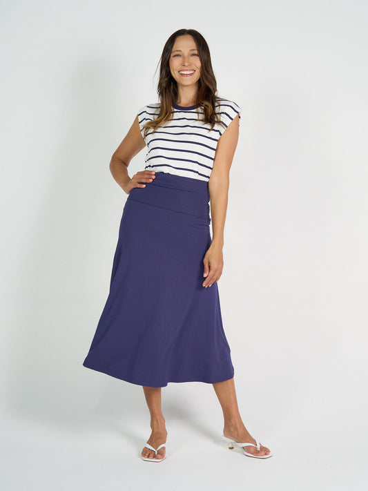 A woman smiles against a white background, wearing the Donna Cap Sleeve Bamboo Tee in blue and white stripes, paired with a high-waisted purple midi skirt and white sandals—an effortlessly chic everyday look.