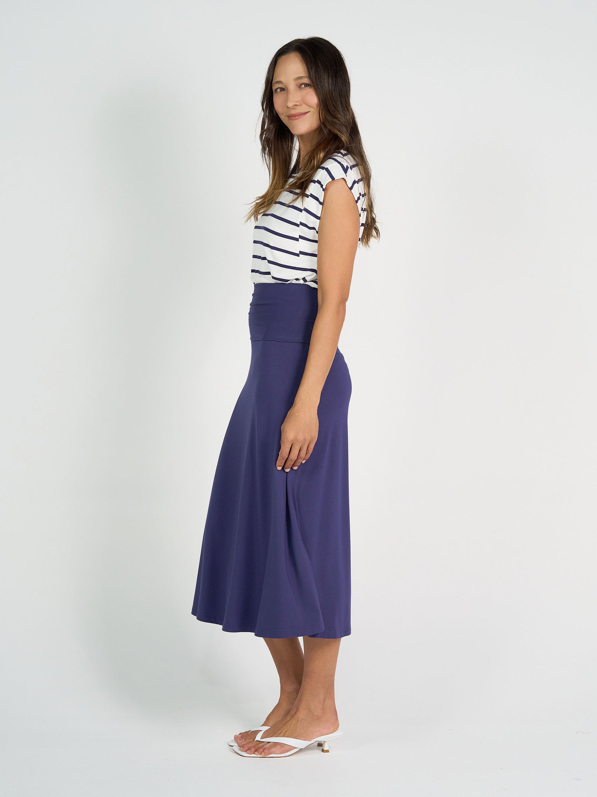 A woman stands sideways in everyday wear: the Donna Cap Sleeve Bamboo Tee in blue & white stripe, paired with a high-waisted purple midi skirt and white sandals. She has long, wavy hair and a slight smile against a white background.