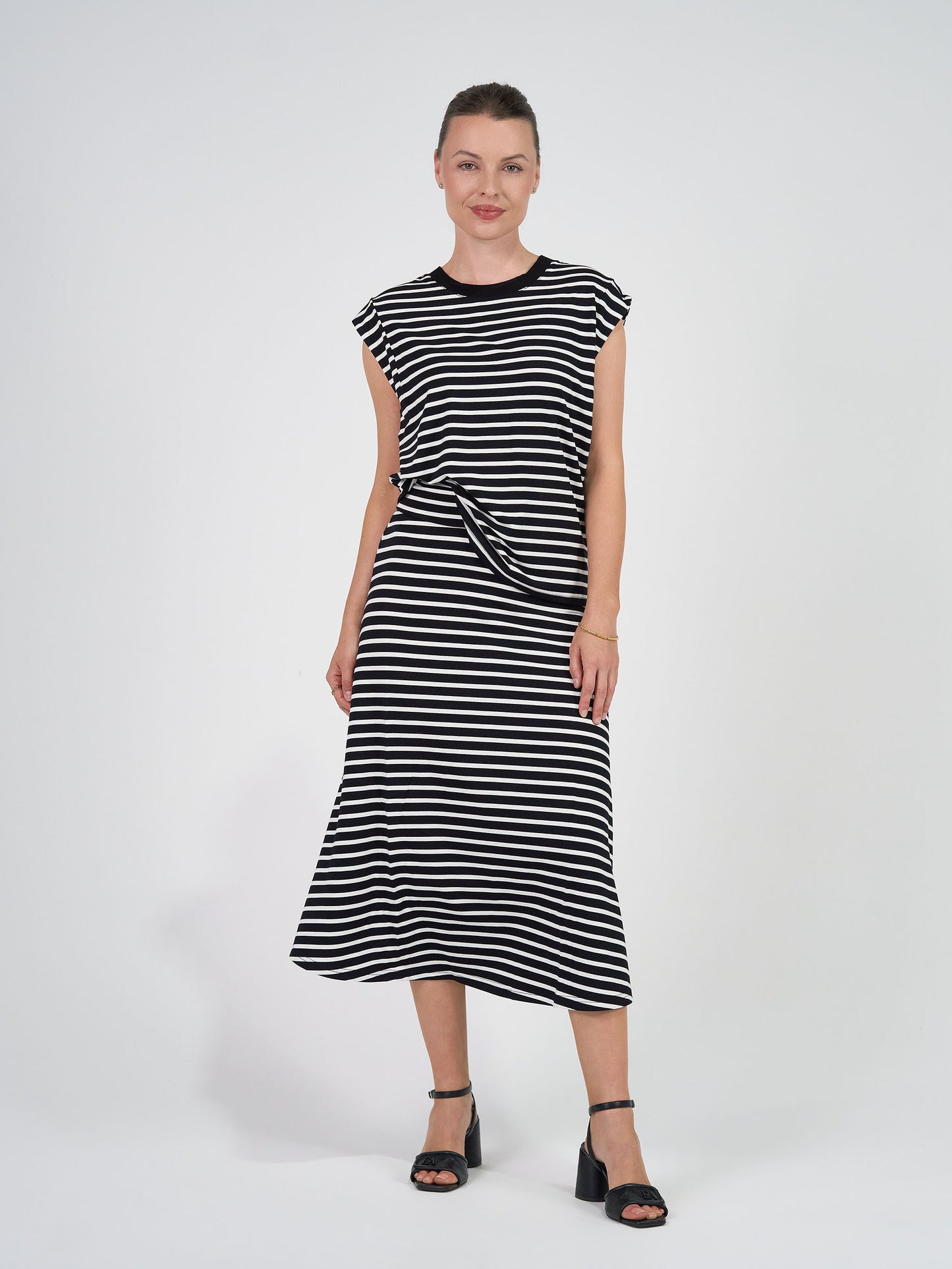 A woman smiles with her hair tied back, wearing the Donna Cap Sleeve Bamboo Tee under a black and white striped midi dress, paired with black heeled sandals against a white background.