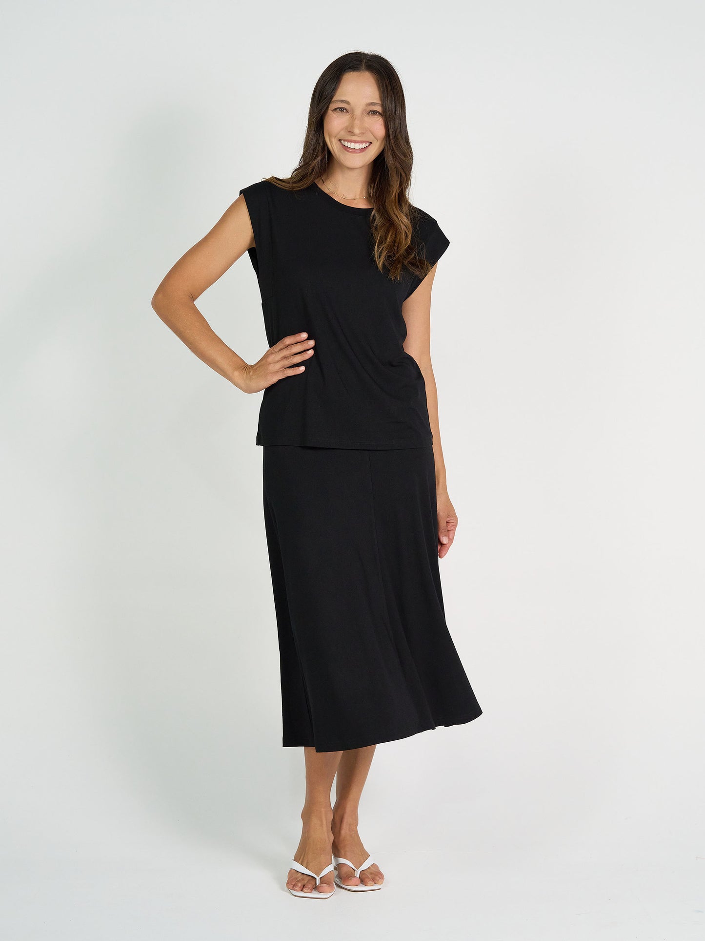 A woman with long brown hair smiles against a white background, wearing the Donna Cap Sleeve Bamboo Tee paired with a black midi skirt and white sandals for an effortlessly stylish everyday look.