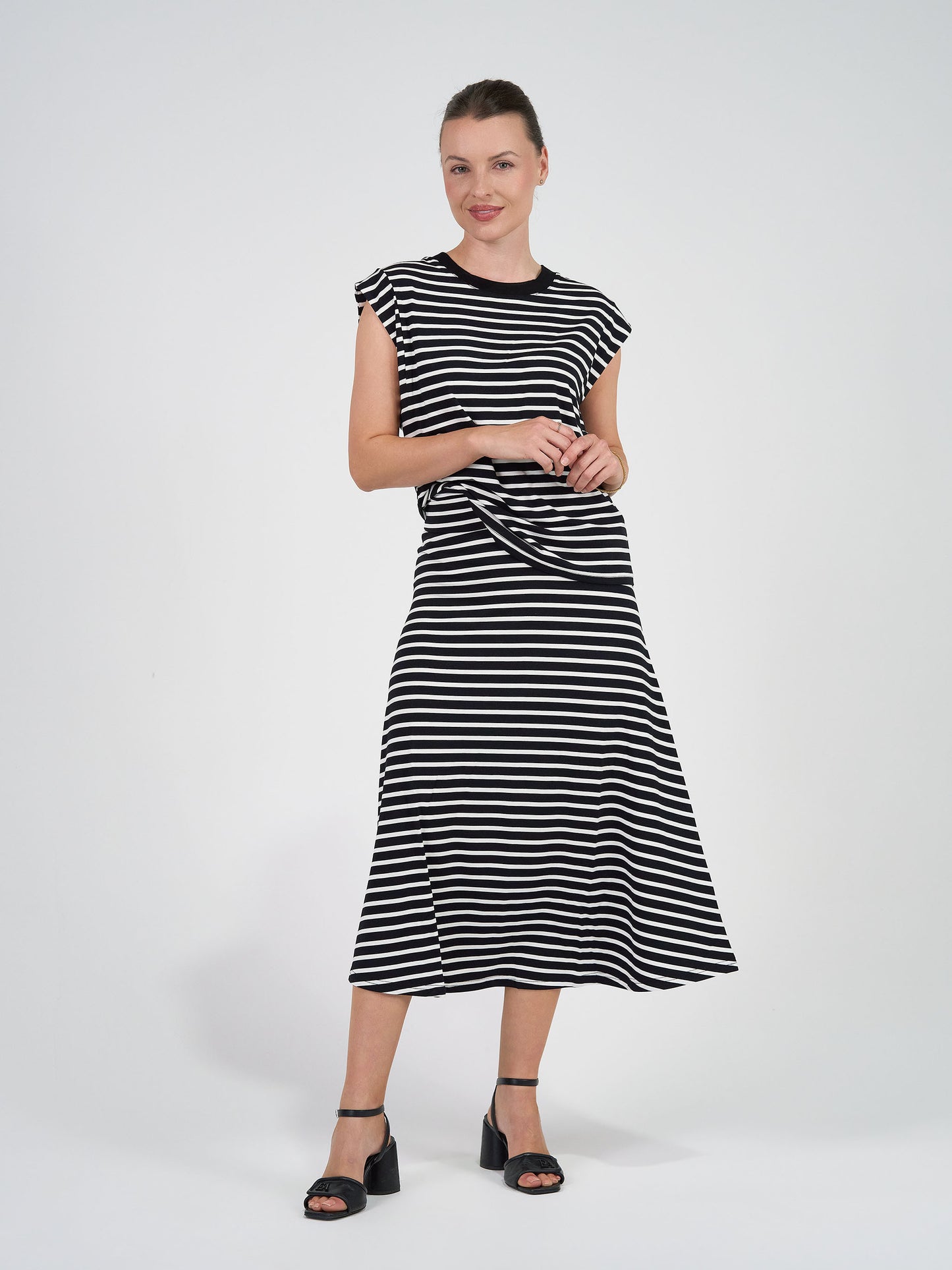 A woman smiles with her hands together, wearing the Jackson Bamboo Maxi Skirt in Black & White Stripe against a plain white background, paired with black heeled sandals.