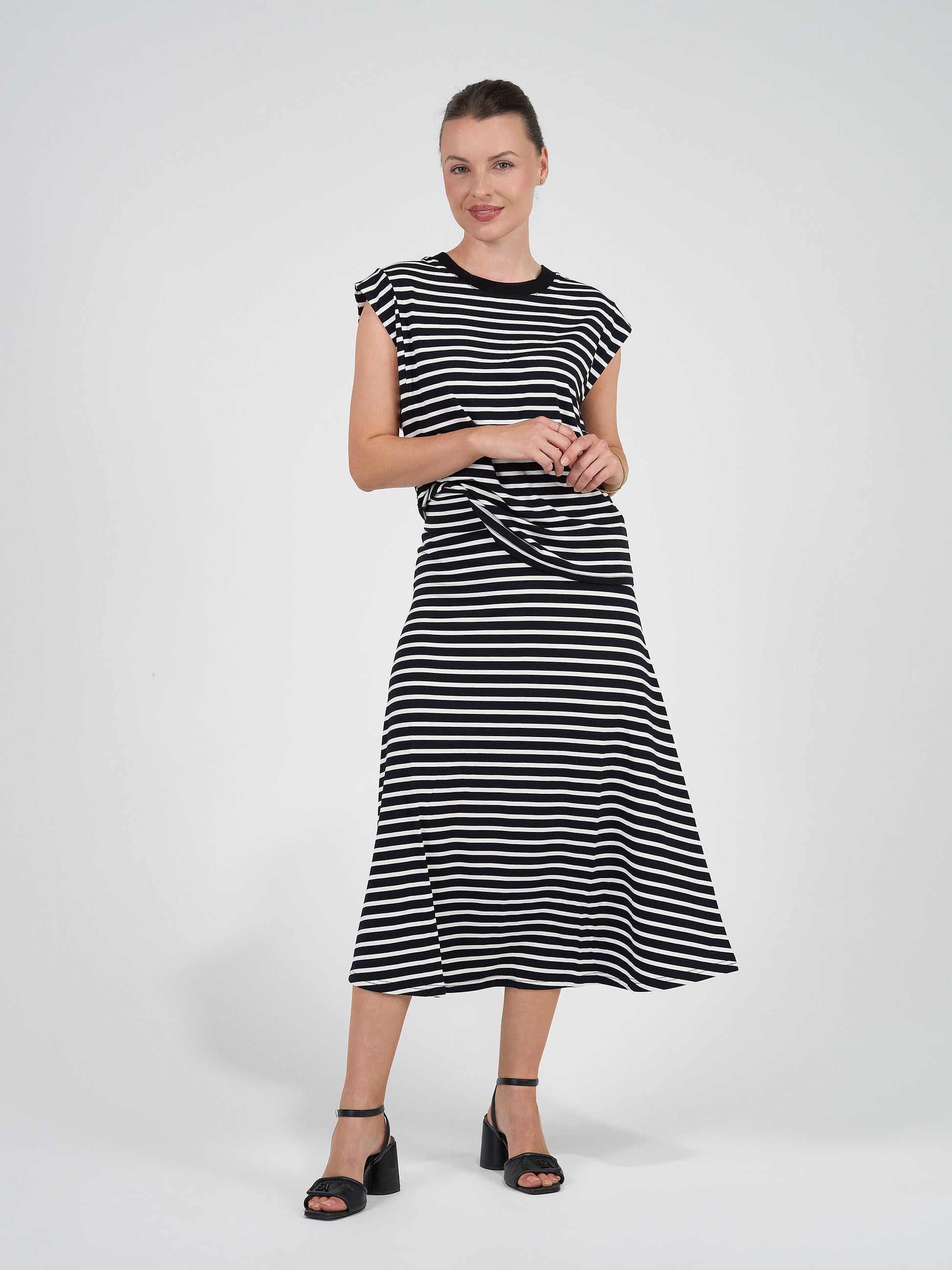 A woman smiles with her hands together, wearing the Jackson Bamboo Maxi Skirt in Black & White Stripe against a plain white background, paired with black heeled sandals.
