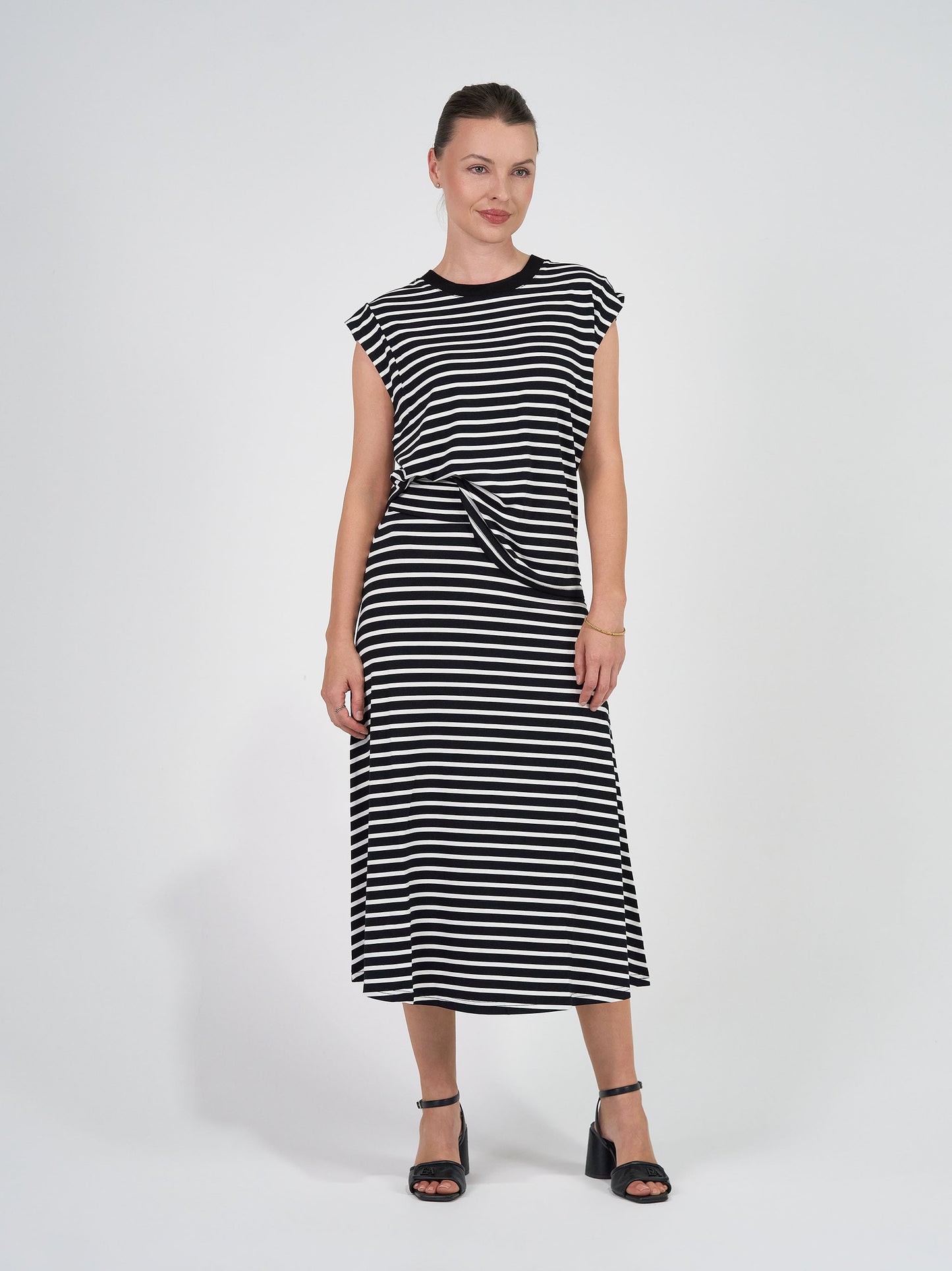 Woman stands against a white background wearing the Donna Cap Sleeve Bamboo Tee in black & white stripe, paired with black heels. Her hair is tied back and she poses with a slight smile, showcasing the tee’s flattering silhouette.