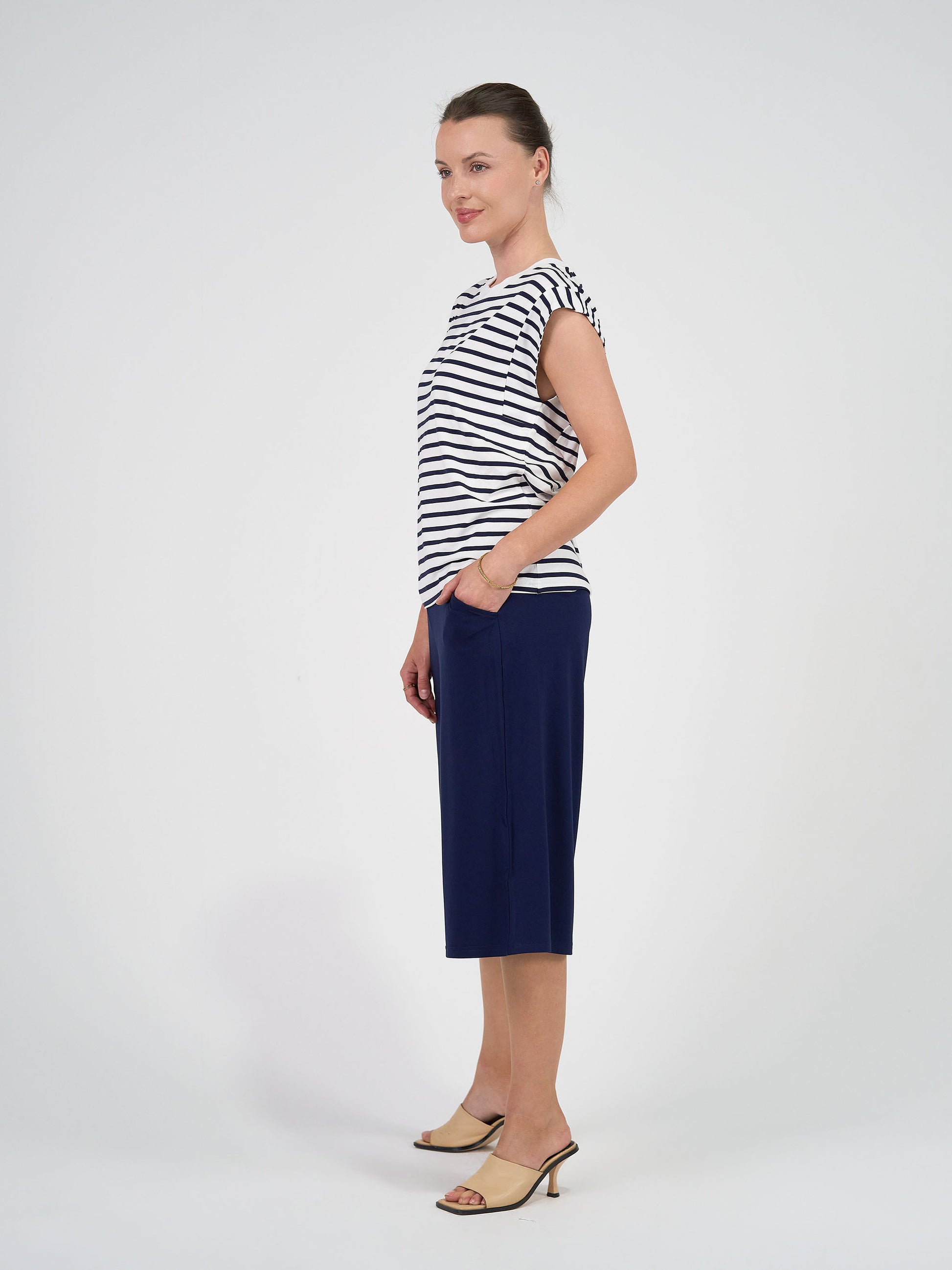 Woman stands sideways against a plain white background, wearing the Donna Cap Sleeve Bamboo Tee in navy and white stripe and a navy skirt, styled with beige heeled sandals. She smiles slightly, hands in pockets—perfect for everyday wear.