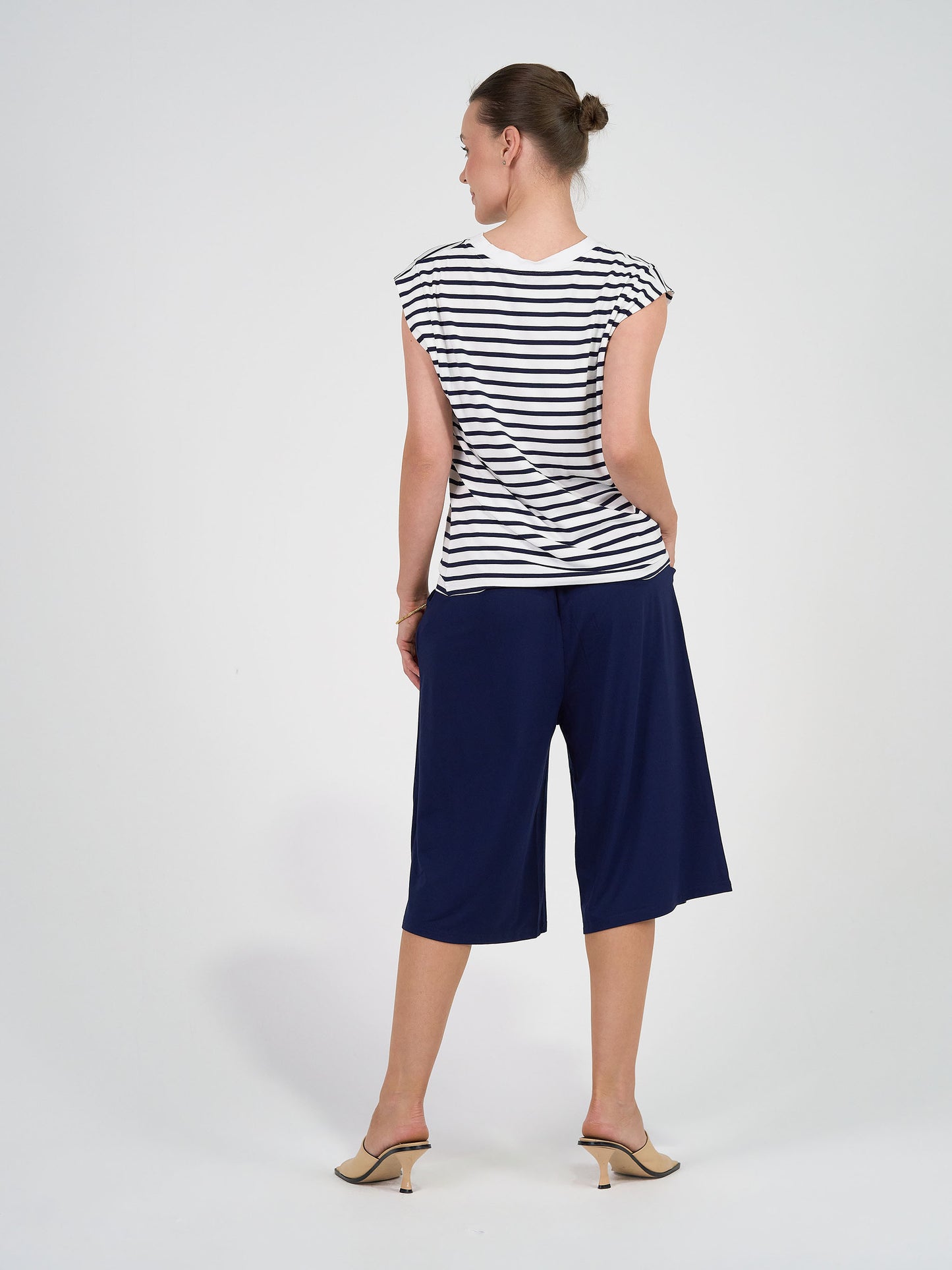 A woman faces away, wearing the Donna Cap Sleeve Bamboo Tee in navy & white stripe, paired with navy culottes and beige heeled sandals. Her hair is in a bun, hands in her pockets, creating a flattering silhouette against a plain white background.