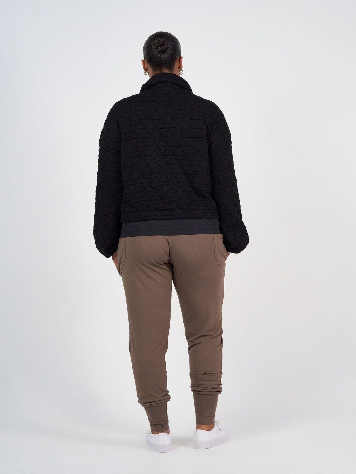 A person stands facing away from the camera, wearing a black Grace Quilted Bamboo Zip Up Jacket, taupe jogger pants, and white sneakers against a plain light background.
