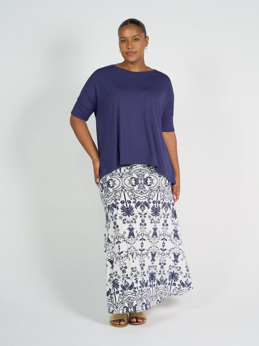 A woman with short hair and a calm expression stands against a white background wearing the Gina Oversized Slouch Bamboo T-Shirt in blue, a long white maxi skirt with blue floral patterns, and sandals.