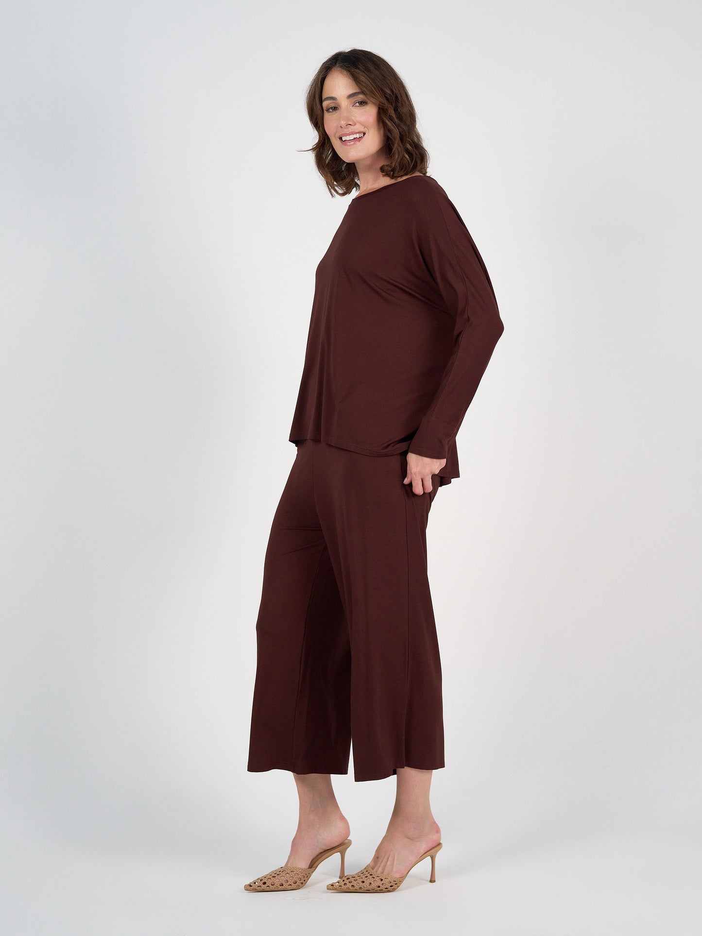A woman with shoulder-length brown hair smiles in the Gina Long Sleeved Bamboo Top in Burgundy and matching wide-leg pants, paired with beige heeled sandals against a plain white background.