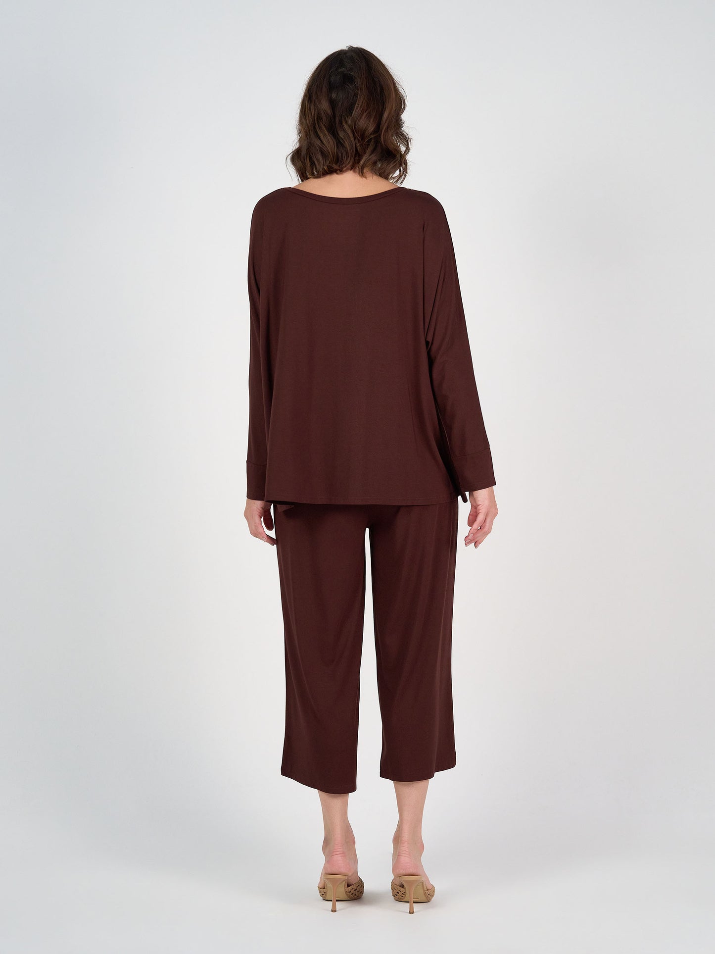 A woman with shoulder-length wavy hair faces away, wearing the Gina Long Sleeved Bamboo Top in burgundy and wide-leg pants with beige heeled sandals. The background is plain white.
