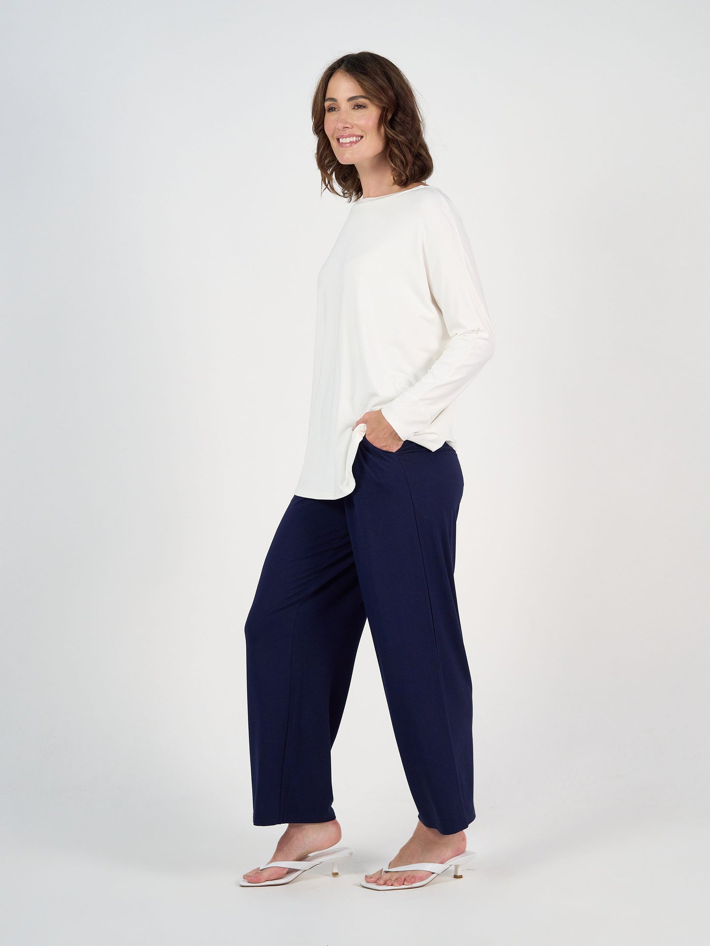 A woman stands against a white background, smiling in the Gina Long Sleeved Bamboo Top - Cream, paired with navy wide-legged pants and white sandals. Her left hand is in her pocket as she looks slightly to the side.