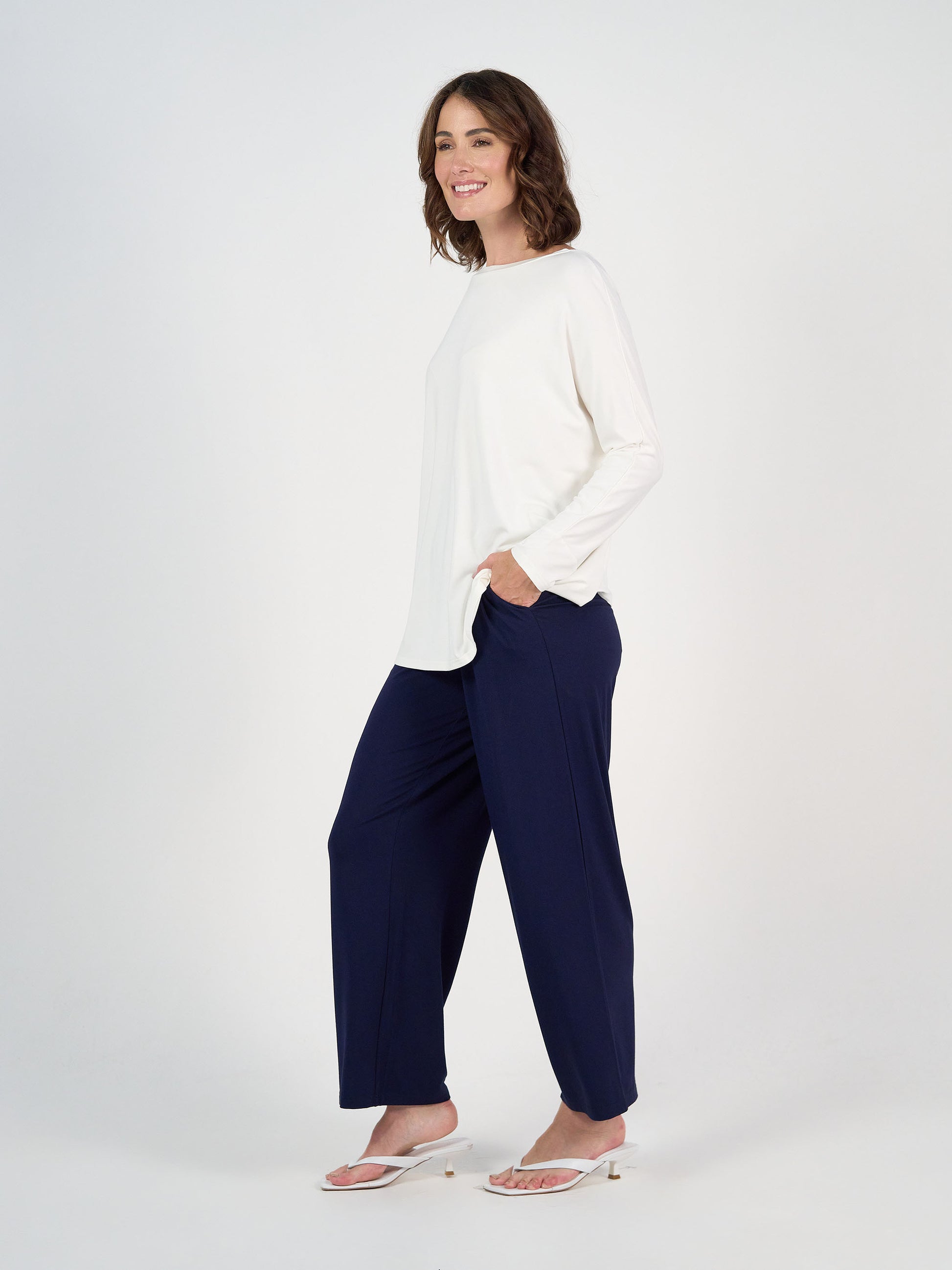 A woman stands against a white background, smiling in the Gina Long Sleeved Bamboo Top - Cream, paired with navy wide-legged pants and white sandals. Her left hand is in her pocket as she looks slightly to the side.