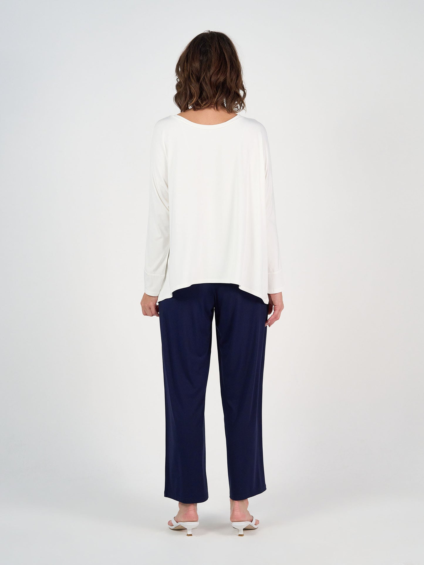 A person with shoulder-length brown hair stands facing away, wearing the Gina Long Sleeved Bamboo Top in cream, navy blue pants, and white sandals against a plain white background.