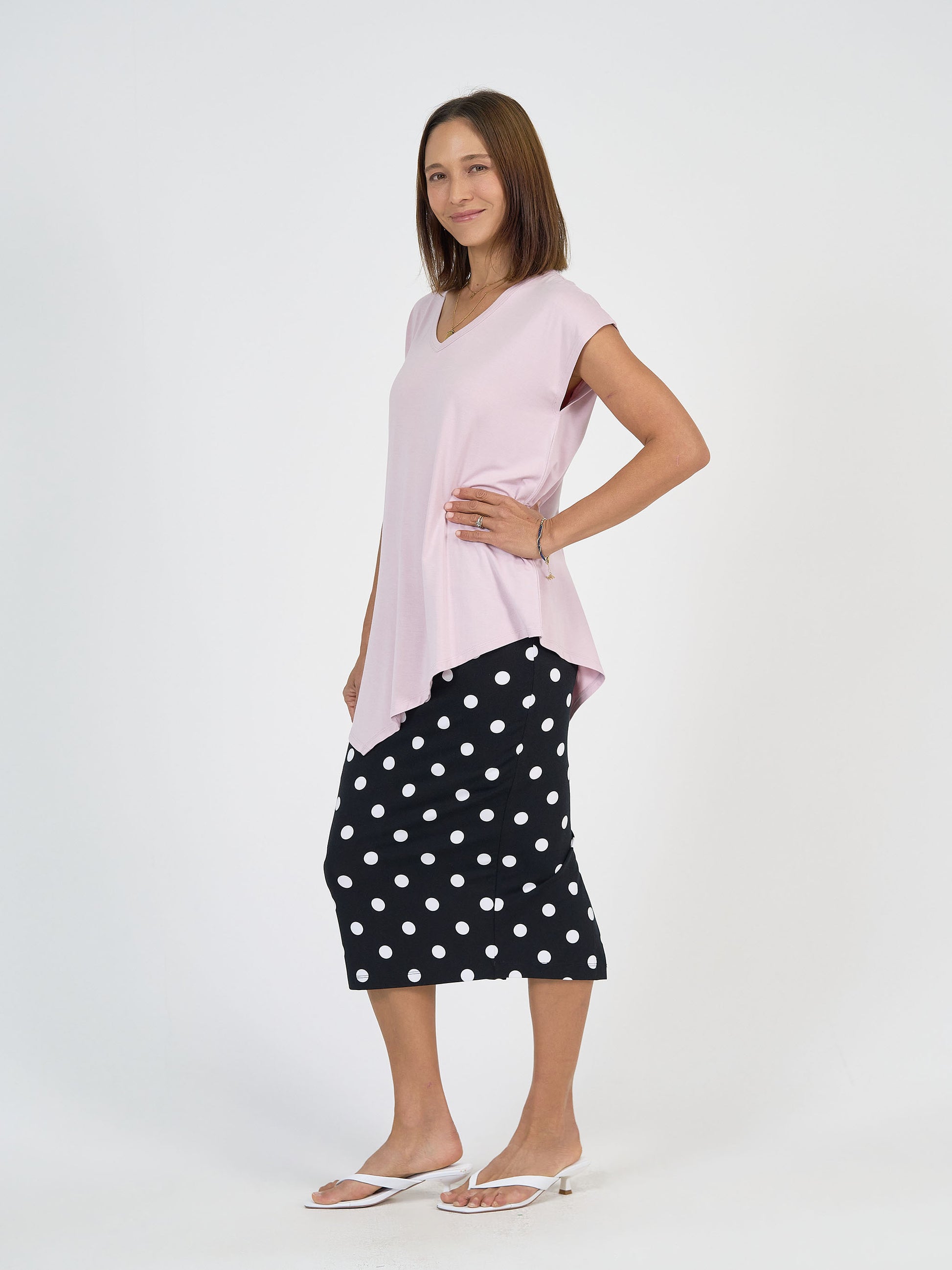 A woman stands and smiles, wearing a light pink asymmetrical top, the Maxi Bamboo Whitney Tube Skirt - Polka Dot in black with white polka dots, white sandals, and a bracelet, against a plain white background.