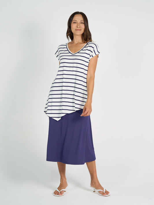 A woman poses casually against a white background, smiling as she wears the Holiday Asymmetrical Bamboo V-Neck T-Shirt in blue & white stripes, paired with a long blue skirt and white sandals.