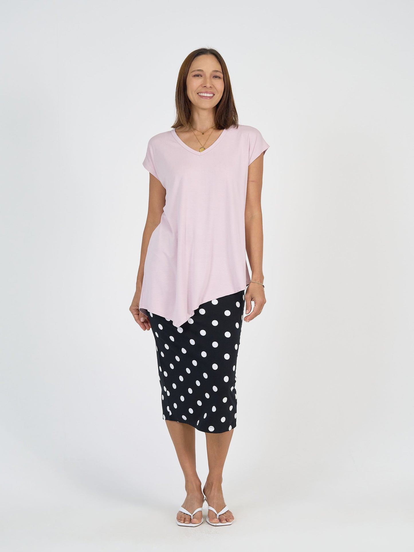 A woman smiles in the Holiday Asymmetrical Bamboo V-Neck T-Shirt in lilac, paired with a black polka dot skirt and white sandals, standing against a plain white background.